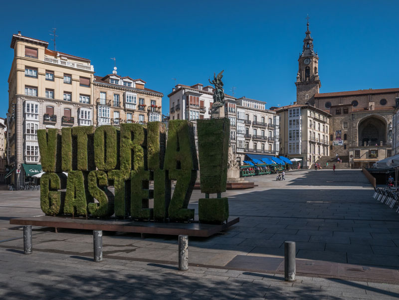 Escultura vegetal con el texto “VITORIA GASTEIZ!” en la plaza de la Virgen Blanca, rodeada de edificios históricos y con la iglesia de San Miguel al fondo.
