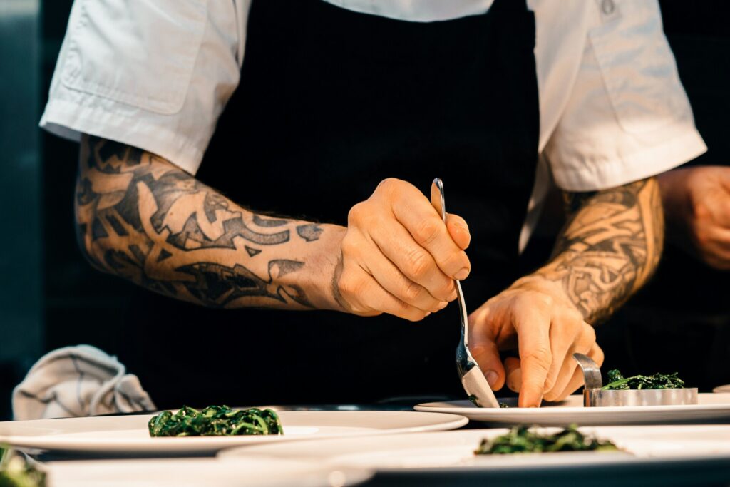 Cocinero con tatuajes preparando un plato en una cocina profesional, colocando verduras con una cuchara.