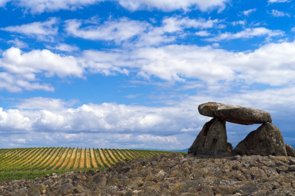 Dolmen de piedra en un paisaje rural con viñedos de la Rioja Alavesa y cielo azul con nubes.